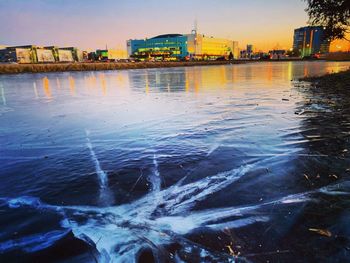 River by illuminated buildings against sky at sunset