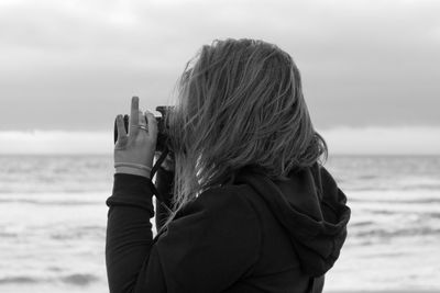 Side view of woman photographing while standing at beach against sky