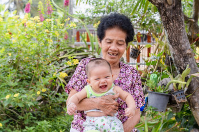 Happy big laughing child boy and senior woman holding adorable baby boy in garden