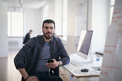 Portrait of male engineer holding mobile phone while sitting at computer desk in office