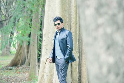 Portrait of confident man with hand in pocket wearing suit and sunglasses while standing against tree in park