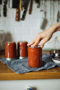 Crop housewife putting lid on glass jar with freshly made tomato sauce while preparing homemade preserves in kitchen
