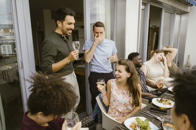 Group of male and female multiracial friends laughing while enjoying drinks at dinner party in balcony