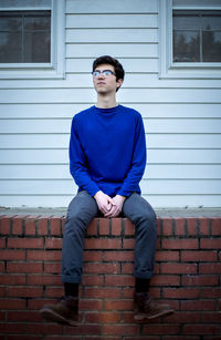 Full length of young man sitting on brick wall against house