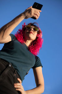 From below of confident female with pink hair surfing cellphone while standing with hand on waist against clear blue sky