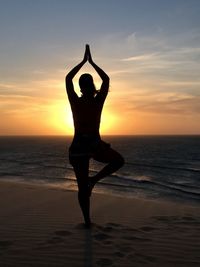 Silhouette woman practicing yoga at beach against sky during sunset