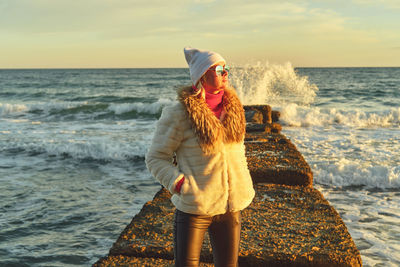 A woman with blond hair in a hat and a fur coat walks along the sea along a stone breakwater