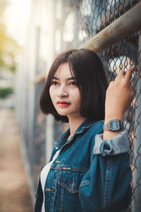 Beautiful young woman looking away while standing by chainlink fence