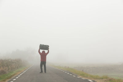 Carrying a suitcase in his arms an unrecognizable man walking along a road on a foggy day