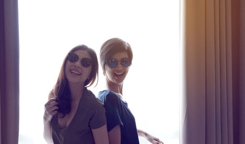 Portrait of smiling lesbians wearing sunglasses against window at home
