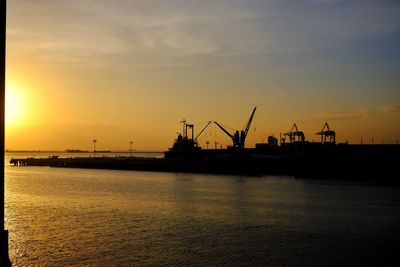 Silhouette cranes at harbor against sky during sunset