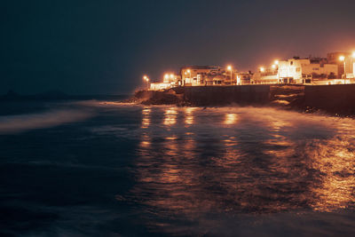 Illuminated buildings by sea against sky at night