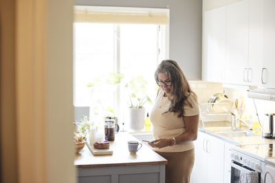 Mature woman using smart phone while standing near kitchen island at home