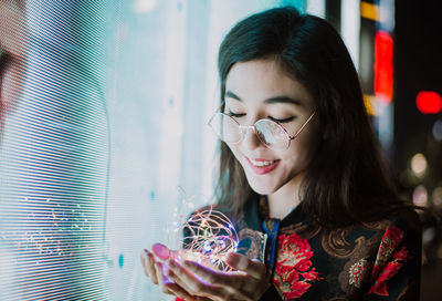 Close-up of smiling young woman holding string lights at night outdoors