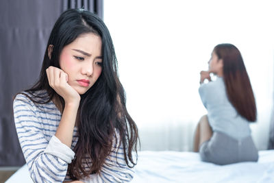 Sad young woman with friend sitting on bed in background