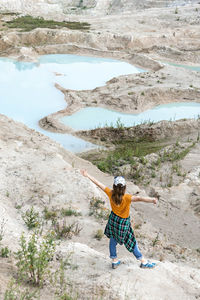 Back view of young woman traveler in cap against  background of blue water of clay quarry and sand 