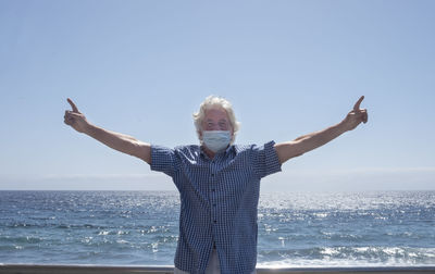 Man wearing mask with arms outstretched standing against sea and clear sky