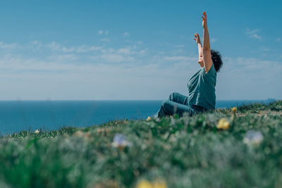 An adult woman sits on a mountain barefoot in a lotus position, raising her hands up to the sky and