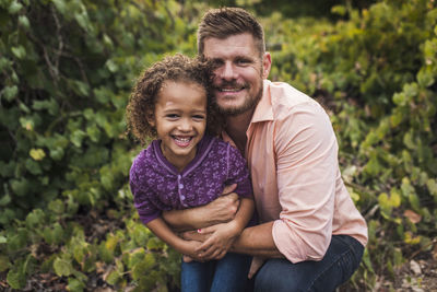 Portrait of father embracing happy daughter while kneeling against plants in park