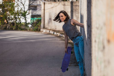 Smiling young woman holding skateboard by road