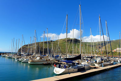 Sailboats moored in sea by mountain against blue sky