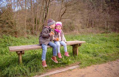 Full length of cute siblings having food while sitting on bench in park