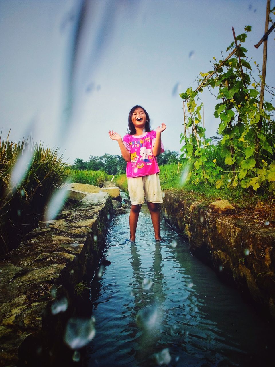 one person, water, nature, sunlight, full length, women, sky, reflection, front view, plant, childhood, child, standing, adult, female, emotion, portrait, flower, clothing, tree, happiness, smiling, leisure activity, outdoors, day, fashion, lifestyles, holiday, casual clothing, young adult, beauty in nature, summer, vacation, trip, looking at camera, pink, land