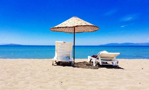 Lounge chairs below parasol at beach against blue sky during sunny day
