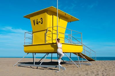 Woman leaning on lifeguard hut at beach against sky