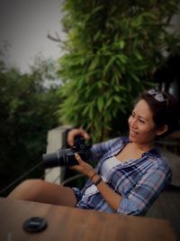 Smiling woman holding dslr camera by table against plants