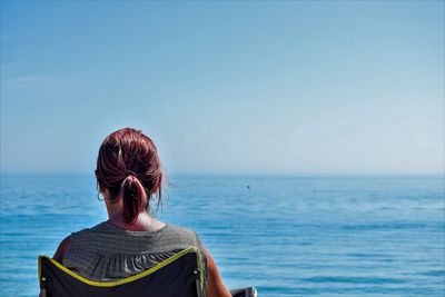 Rear view of mature woman relaxing on chair at beach against sky during sunny day