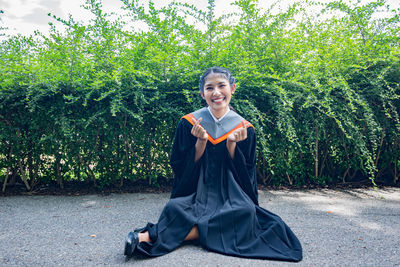 Portrait of smiling young woman wearing graduation gown sitting against plants