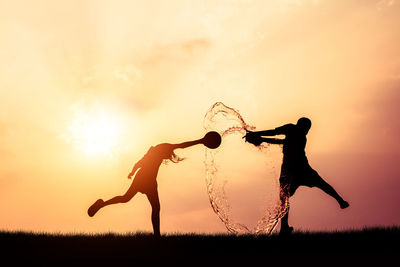 Silhouette friends splashing water with containers while standing on field against orange sky