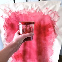 Hand holding mason jar half full of pink paint in front of a painted canva