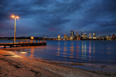 Illuminated buildings by sea against sky at night