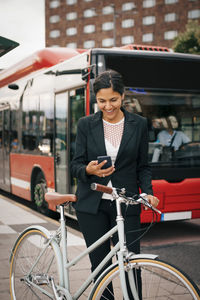Smiling businesswoman using smart phone while standing with bicycle against bus on street in city