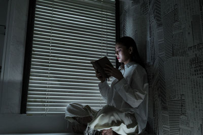 Side view of young woman using mobile phone while sitting on window