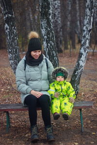 Full length of woman and daughter sitting on bench against tree trunk