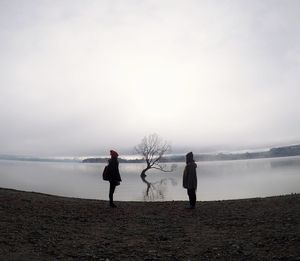 People standing by bare tree at beach against sky during foggy weather