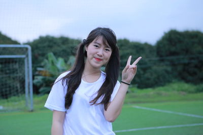 Portrait of smiling young woman gesturing peace sign while standing on playing field