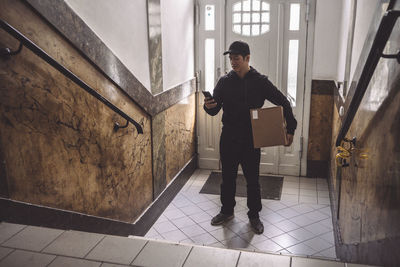 Delivery man using smart phone while holding package against door