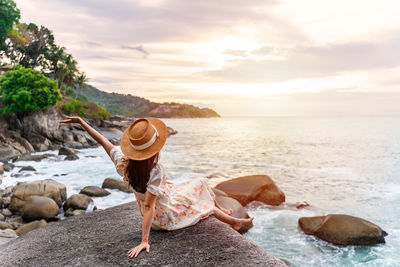 Rear view of woman sitting on rock at beach against sky