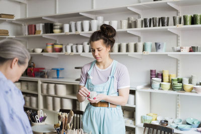 Young female potter holding clay with senior colleague against vases on shelf at workshop