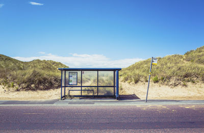 Empty road by sand dunes against blue sky, a bus stop is in the foreground. 