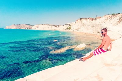 Shirtless man sitting on rock by sea at scala dei turchi during sunny day