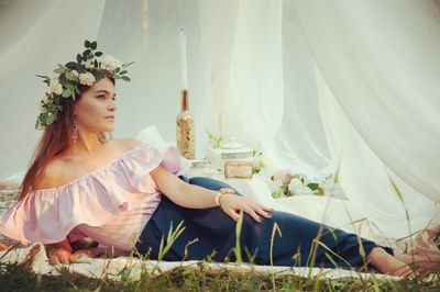 Thoughtful woman wearing wreath resting on field by tent in park