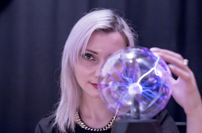 Close-up portrait of young woman holding plasma ball over black background