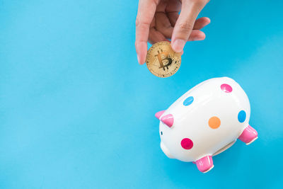 Close-up of hand holding bitcoin by piggy bank against blue background