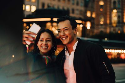 Smiling man and woman taking selfie through phone while standing in city at night