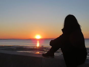 Woman sitting on retaining wall by sea during sunset